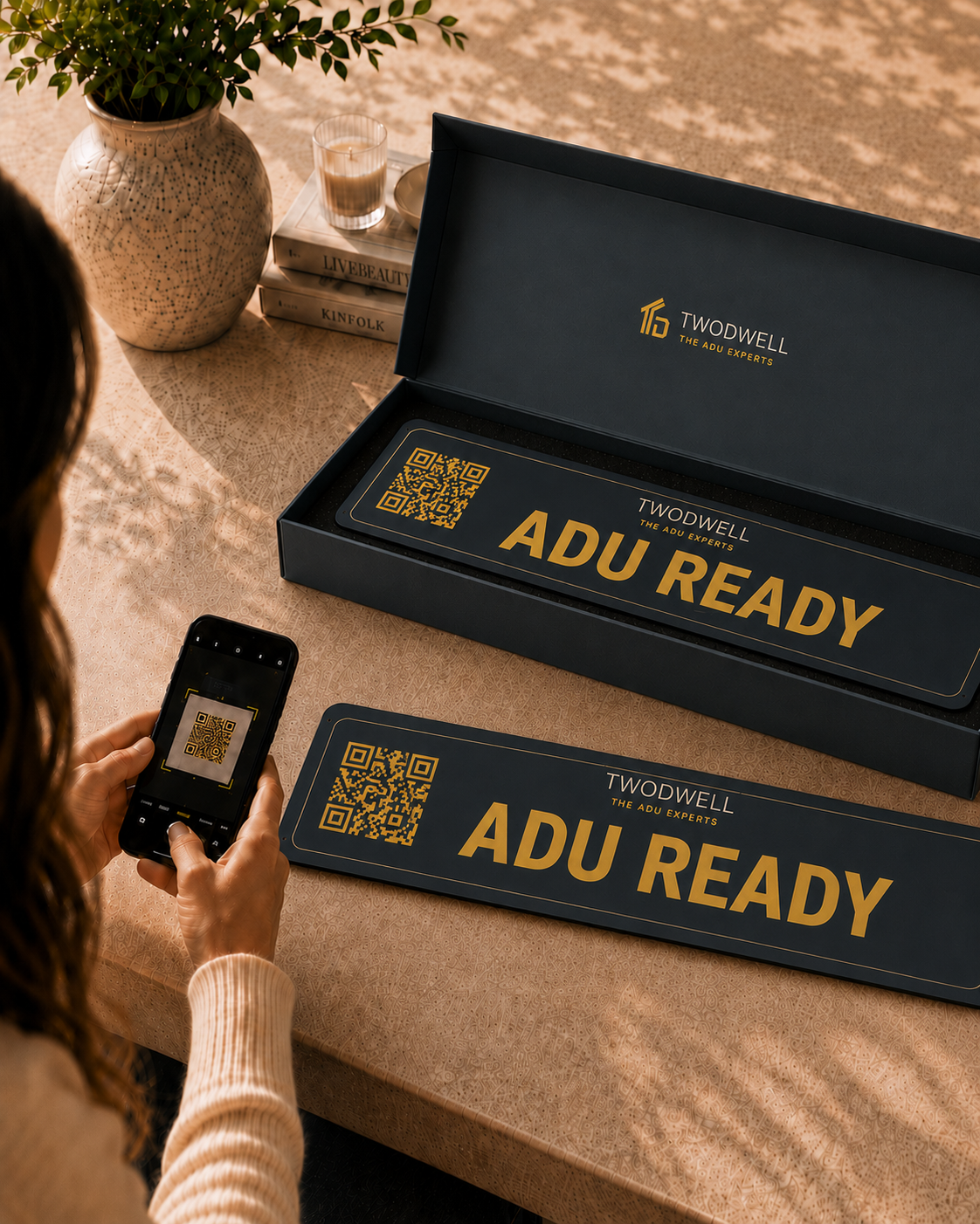 A woman scanning the QR code on a branded TWODWELL ADU Ready rider sign with her phone, next to the premium rider kit box